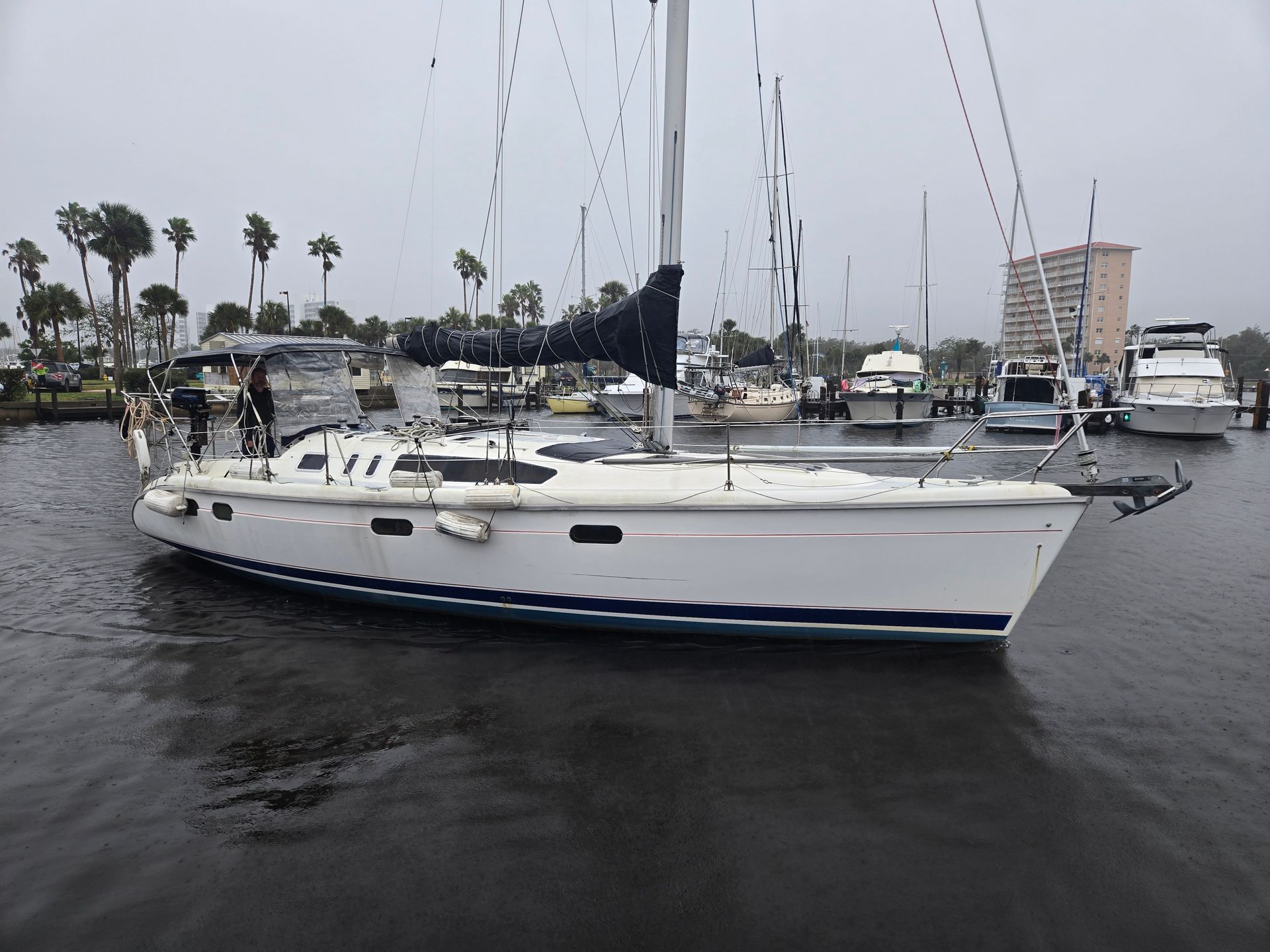 White sailboat on calm water in a marina with palm trees and cloudy sky