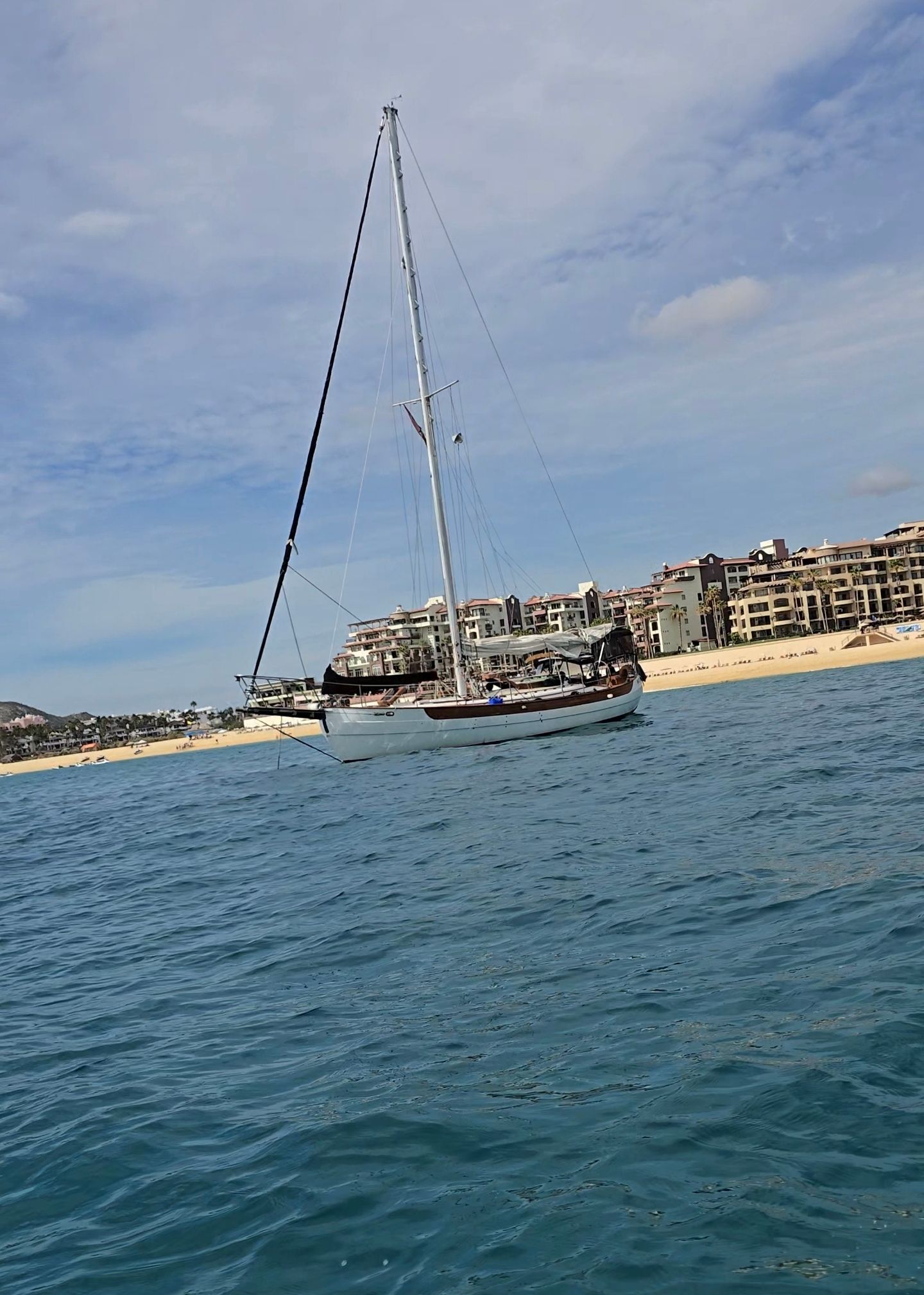 Sailboat on blue water near a sandy beach and waterfront buildings under a partly cloudy sky