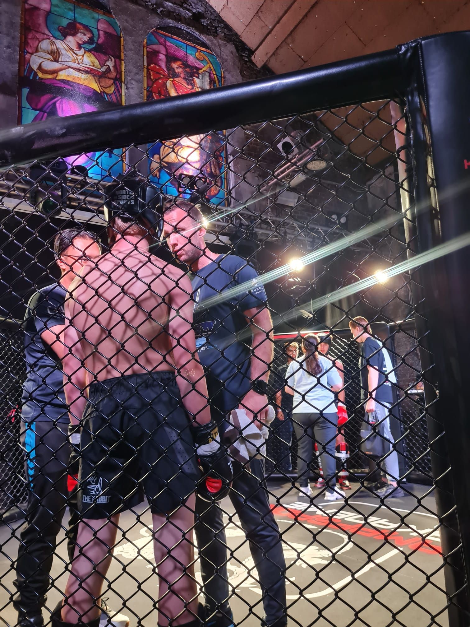 Twee mannen in een MMA-kooi, de ene met ontbloot bovenlijf en de andere in een blauw shirt, met toeschouwers op de achtergrond.