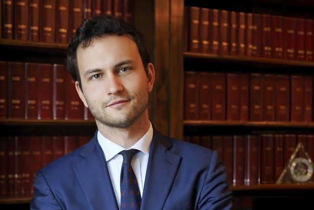 Ritratto di un uomo in abito blu in piedi davanti a degli scaffali in un ufficio o in una biblioteca.