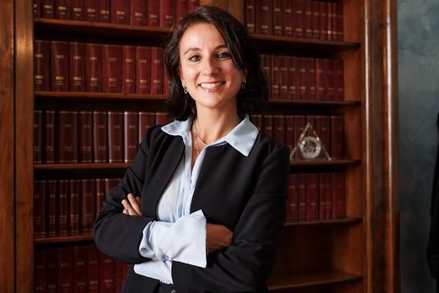 Una donna sorridente in tailleur nero è in piedi davanti a degli scaffali di legno in una biblioteca.