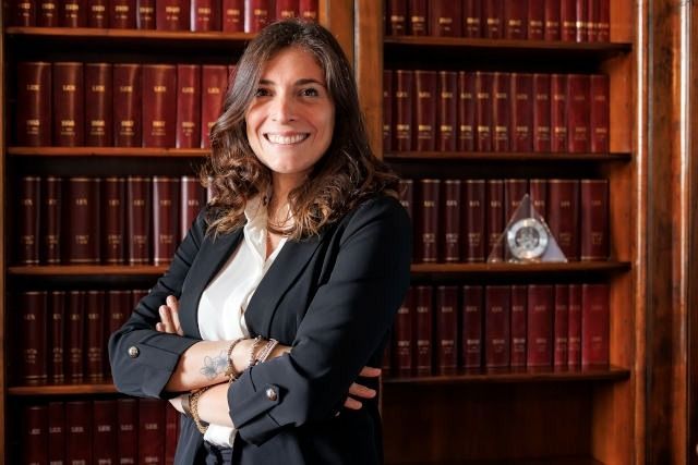 Una persona sorridente con indosso un blazer scuro è in piedi con le braccia incrociate davanti a una libreria di legno.