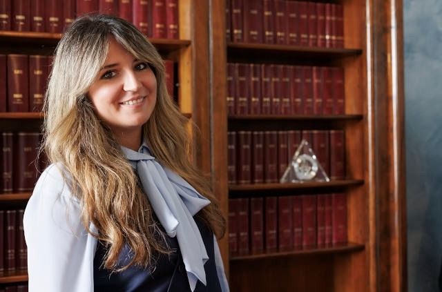Una donna sorride in una biblioteca, in piedi davanti a scaffali di legno pieni di libri.