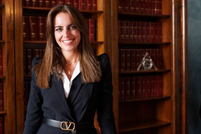 Una donna sorridente in tailleur nero è in piedi davanti a degli scaffali di legno in una biblioteca.