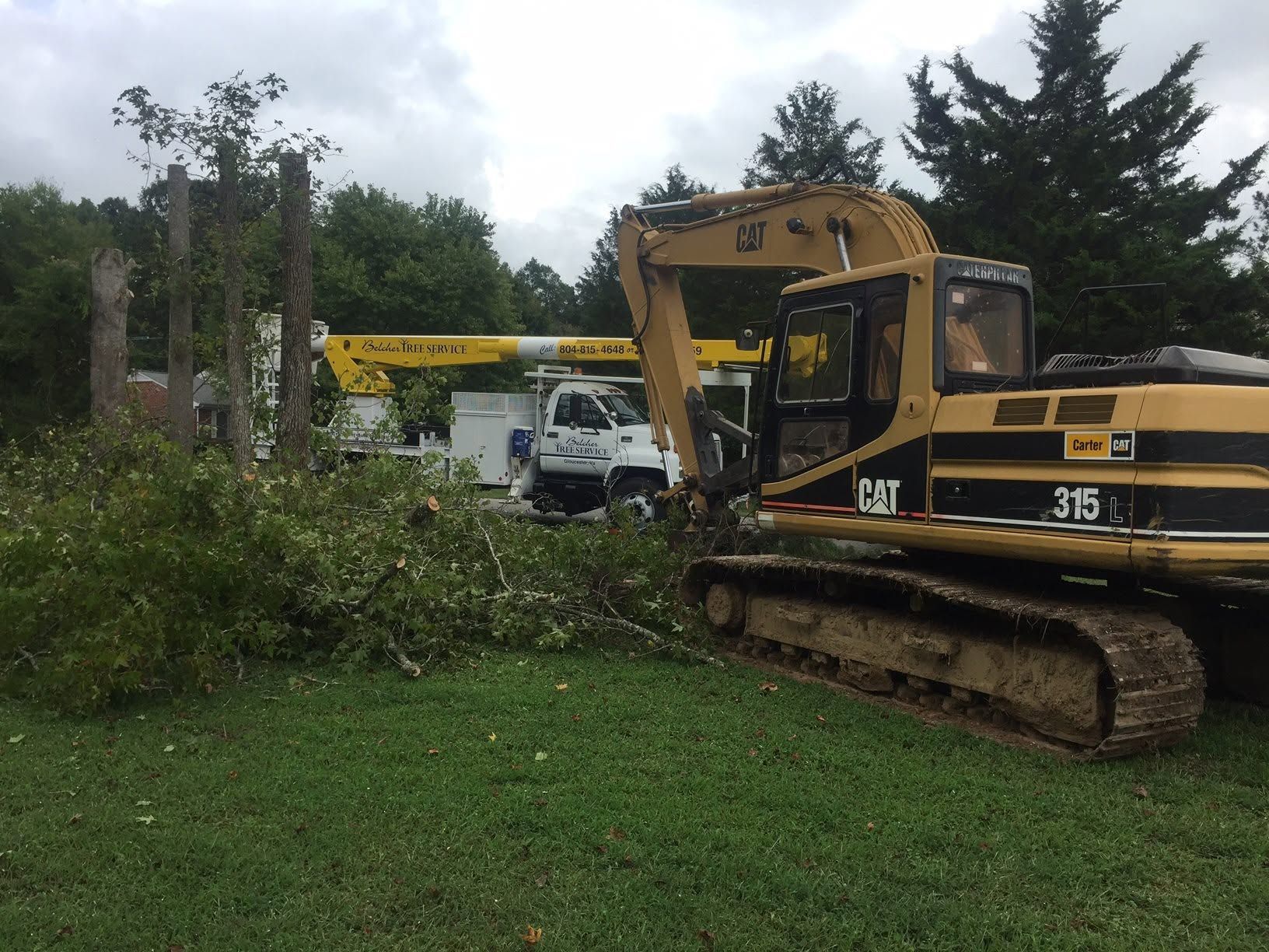 Yellow CAT excavator clearing fallen branches in a grassy yard near a white truck and crane.