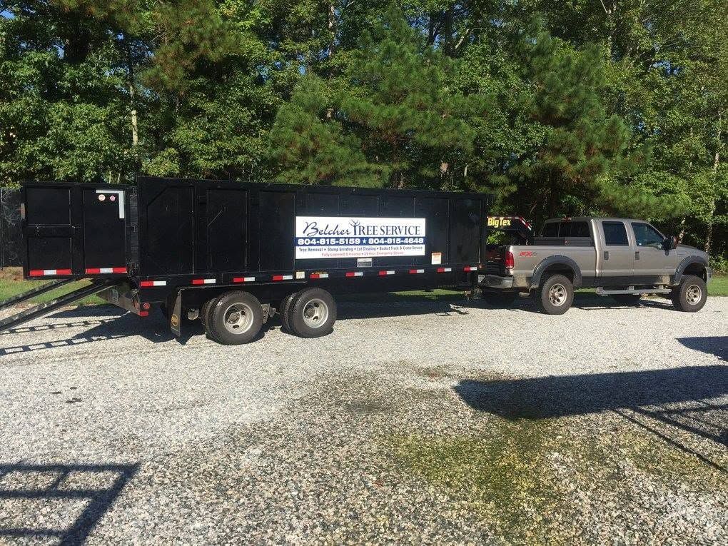 Gray pickup truck towing a black equipment trailer on a gravel driveway, with trees in the background.