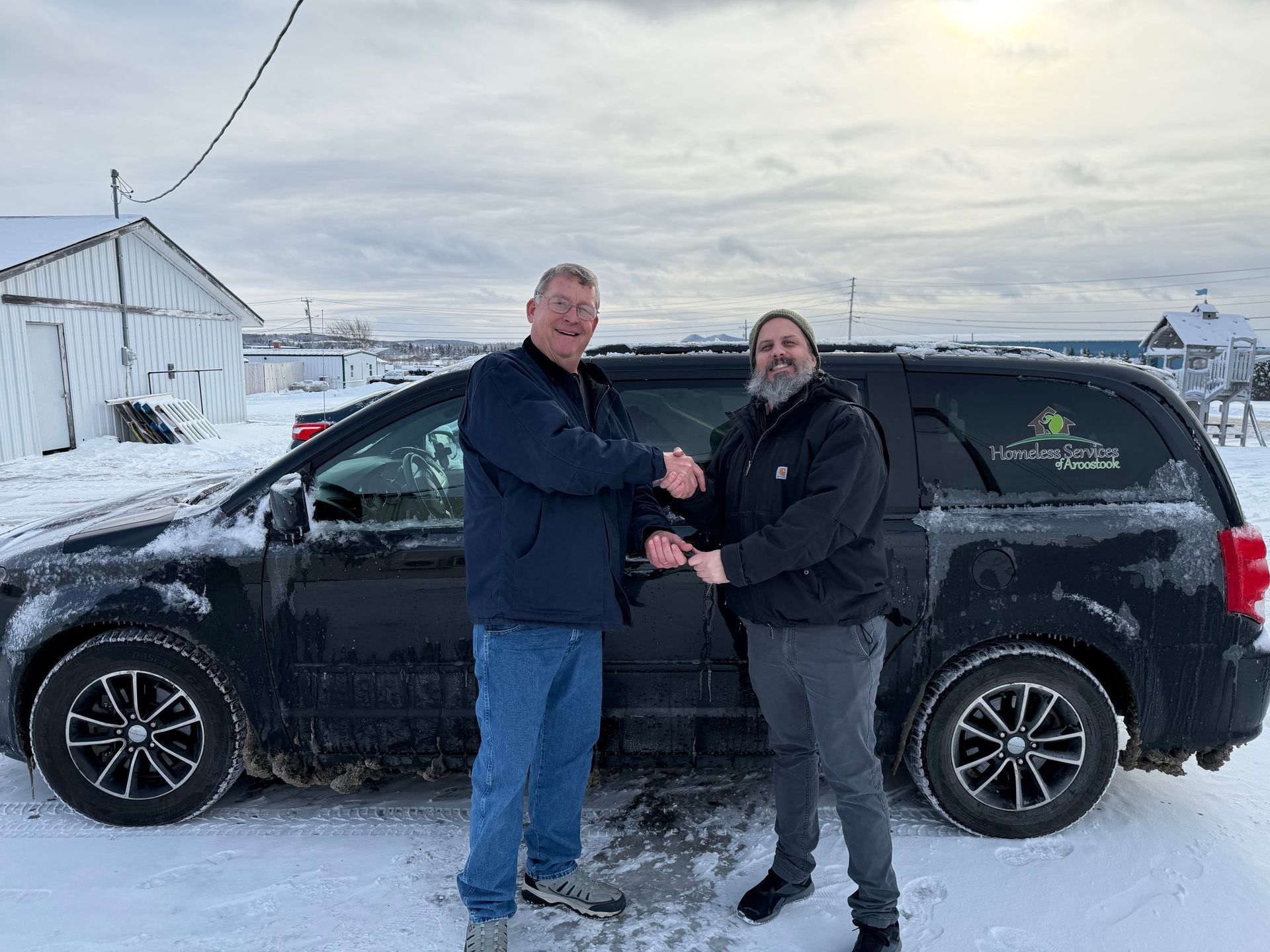 Two people shake hands in front of a black minivan parked on a snow-covered lot on a bright, cloudy day.