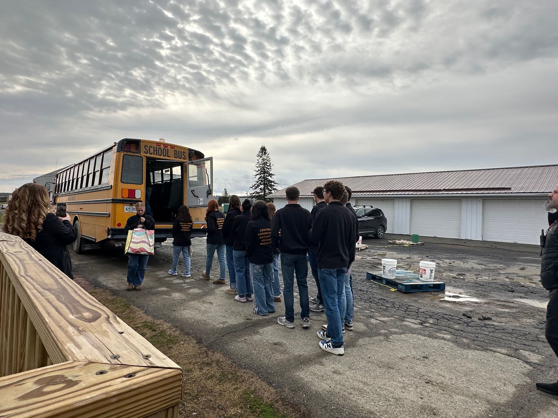 A group of people standing in a line beside a yellow school bus parked in a gravel lot near a long, white building.