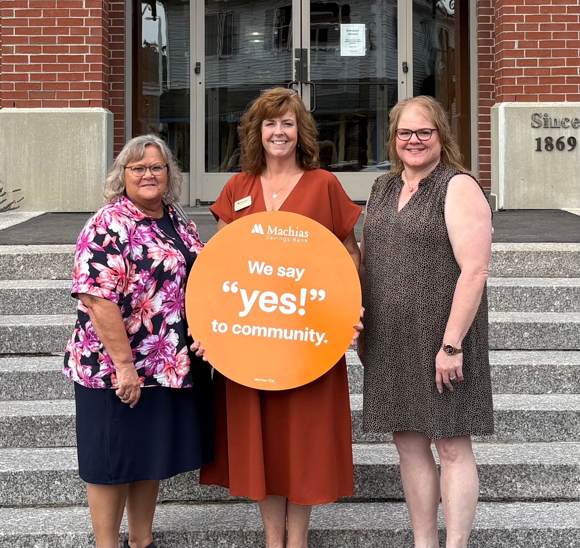 Three people stand on steps in front of a brick building, holding an orange sign that says
