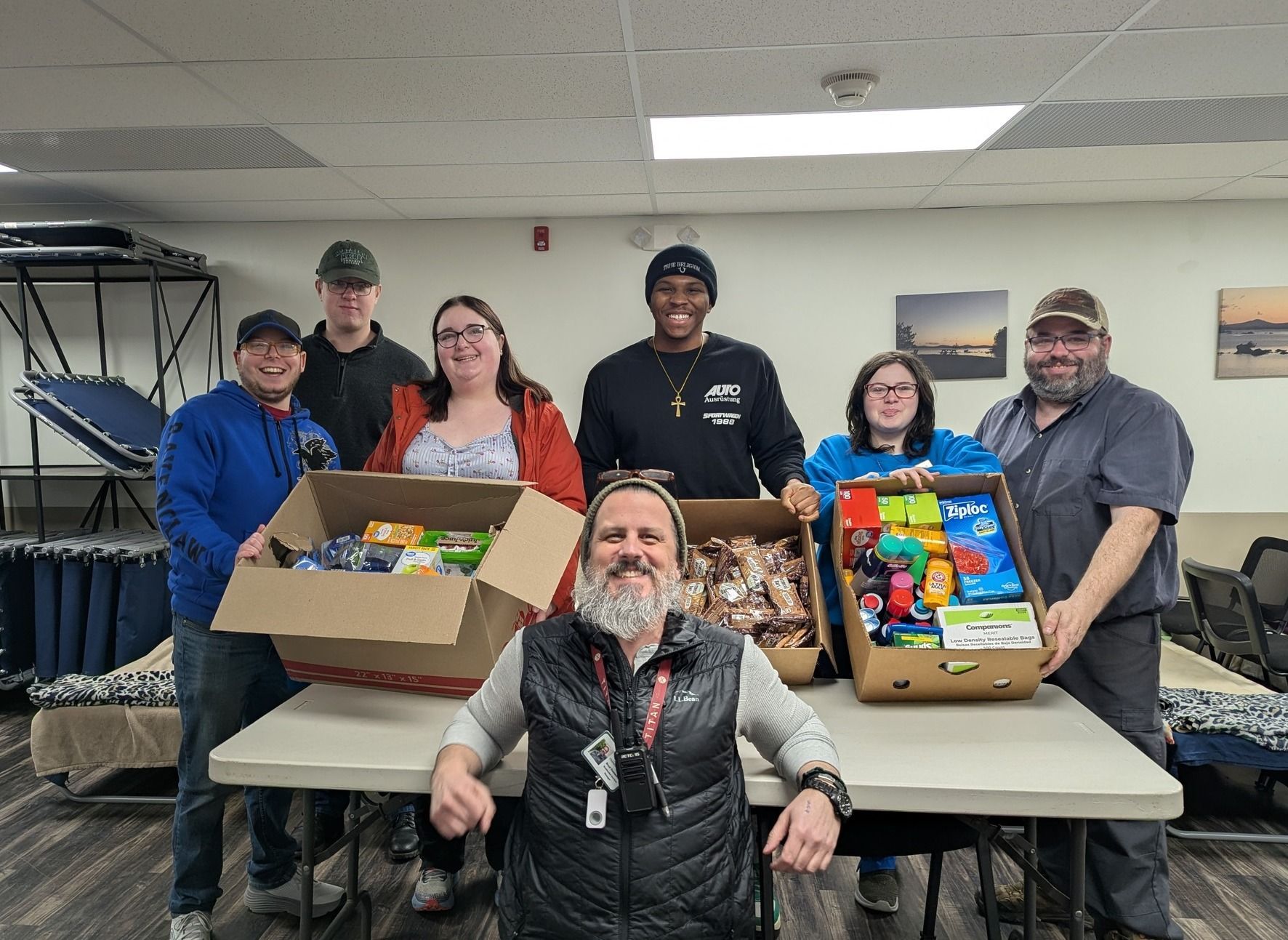 A group of people standing behind boxes of food and supplies for a donation drive in an indoor entryway.