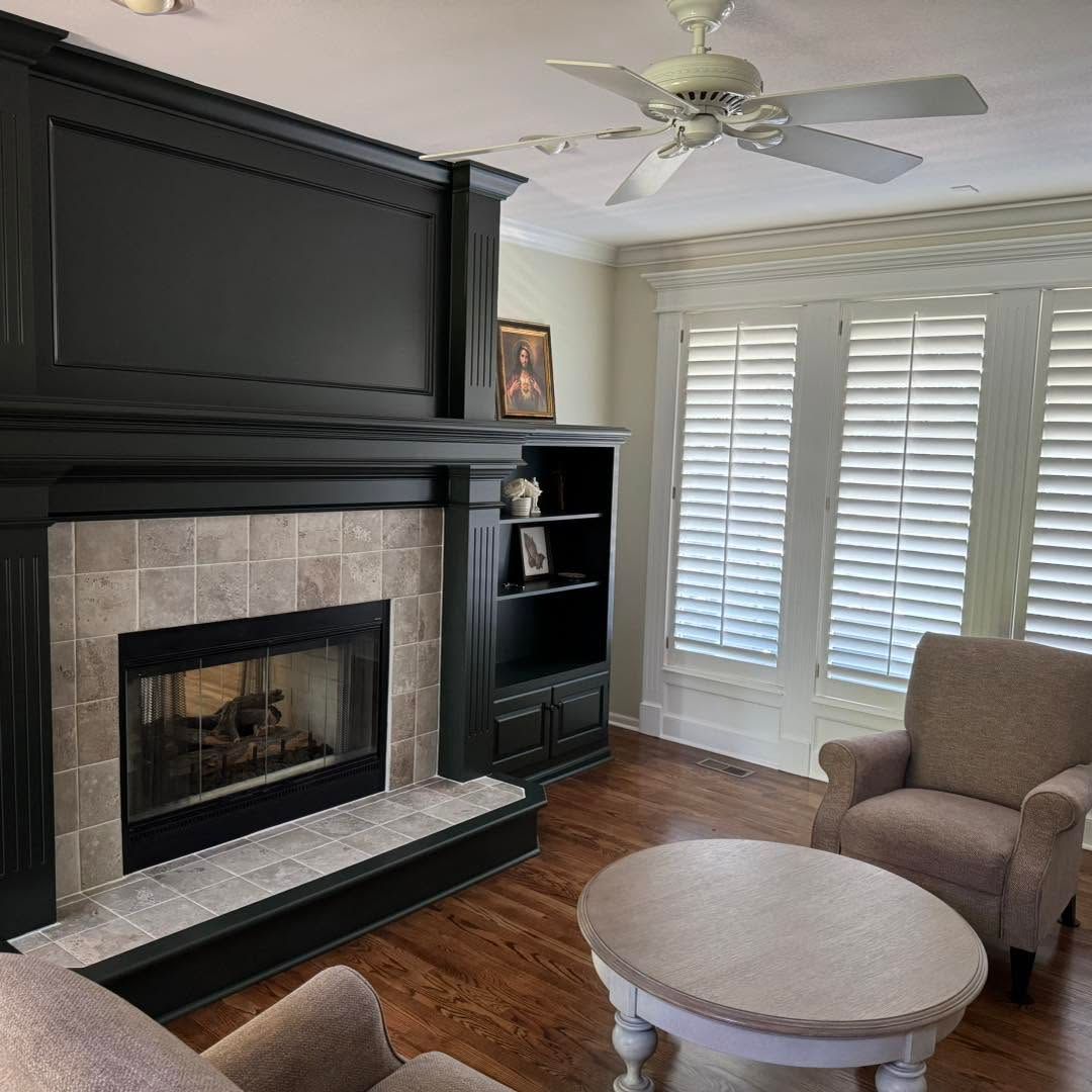 Bright living room with a black fireplace, white shutters, ceiling fan, and beige chair by a round table