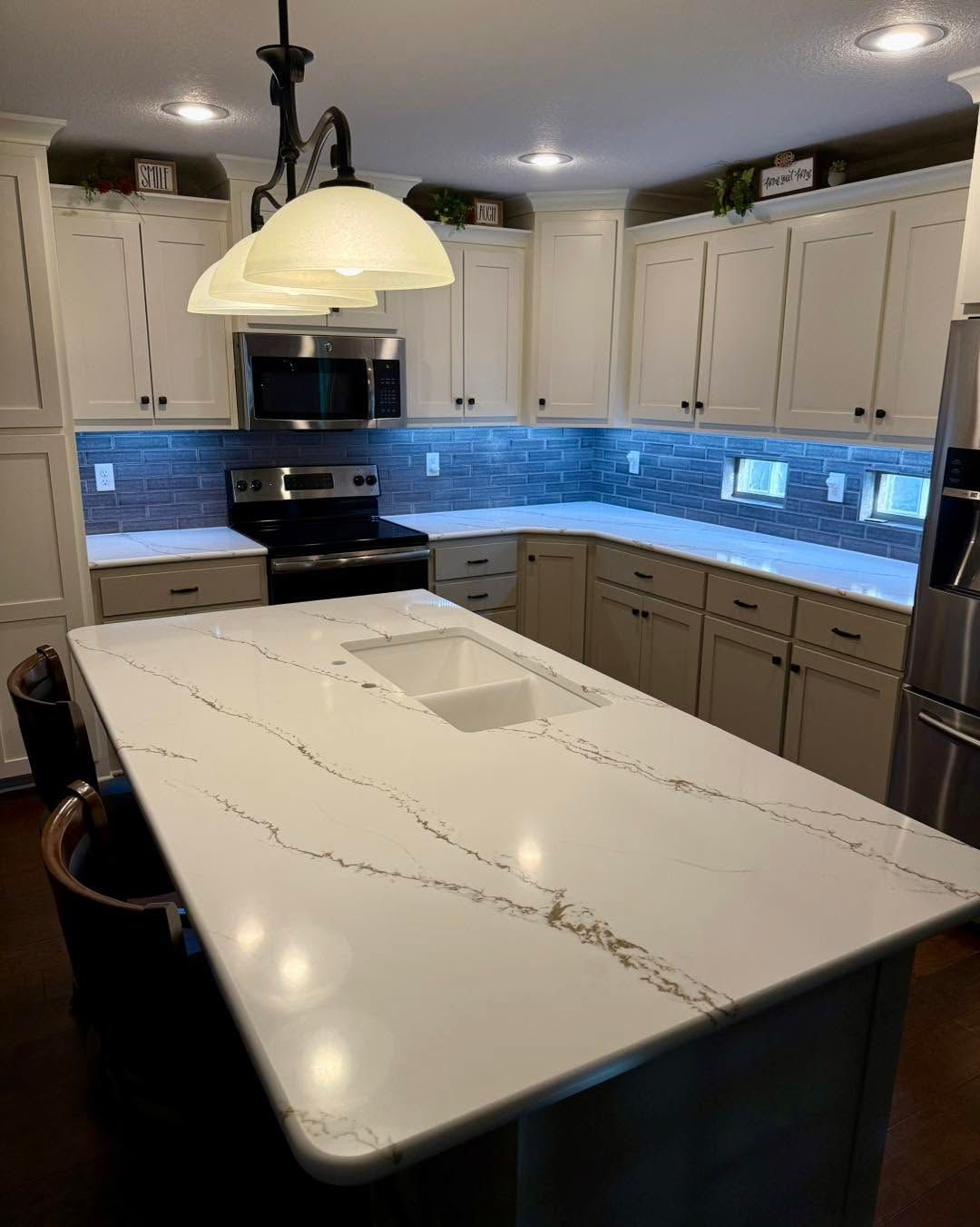 Modern kitchen with white island, gray cabinets, blue backsplash lighting, and hanging pendant lights.