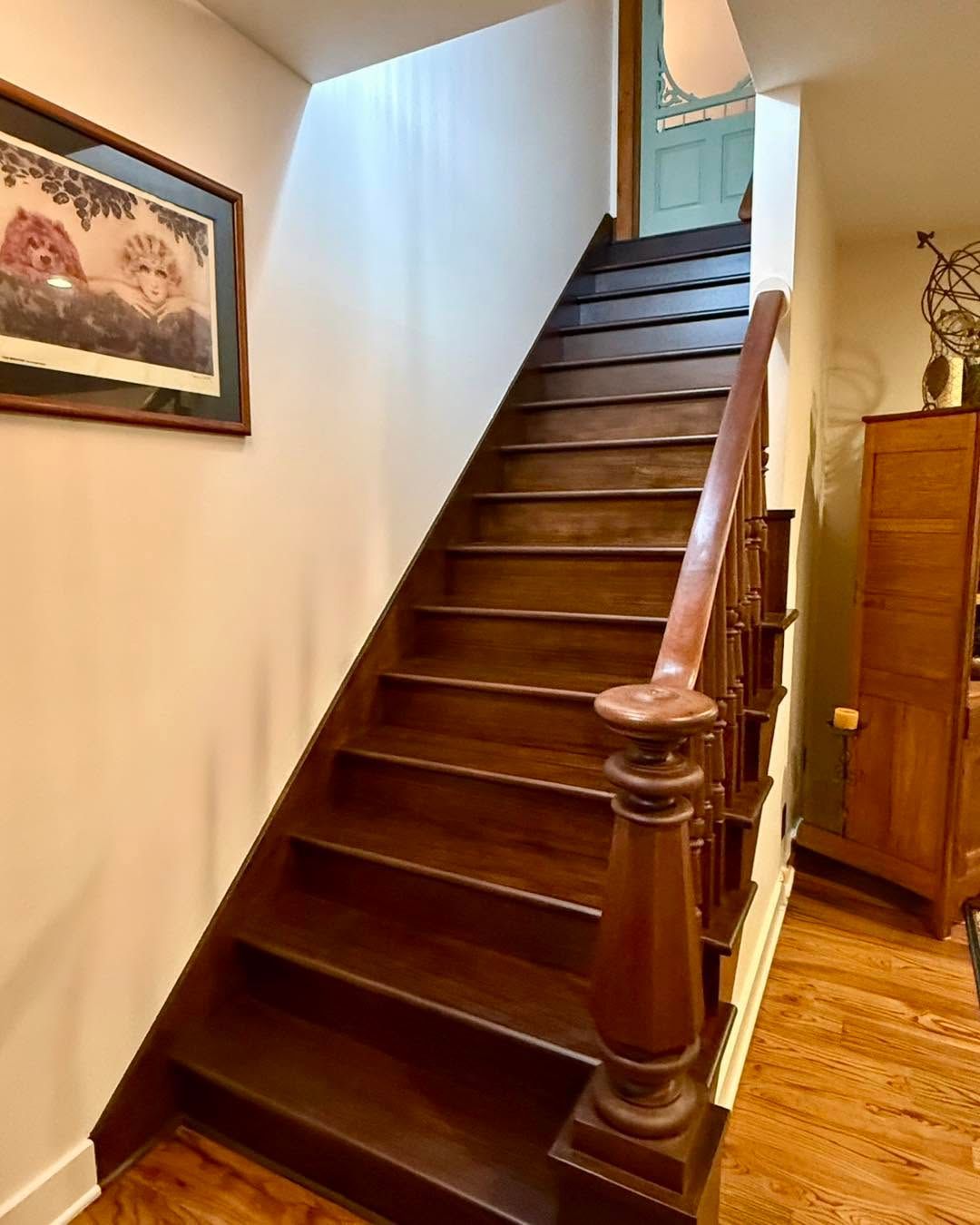 Wooden staircase with dark railings in a warmly lit home interior, beside a framed wall picture.