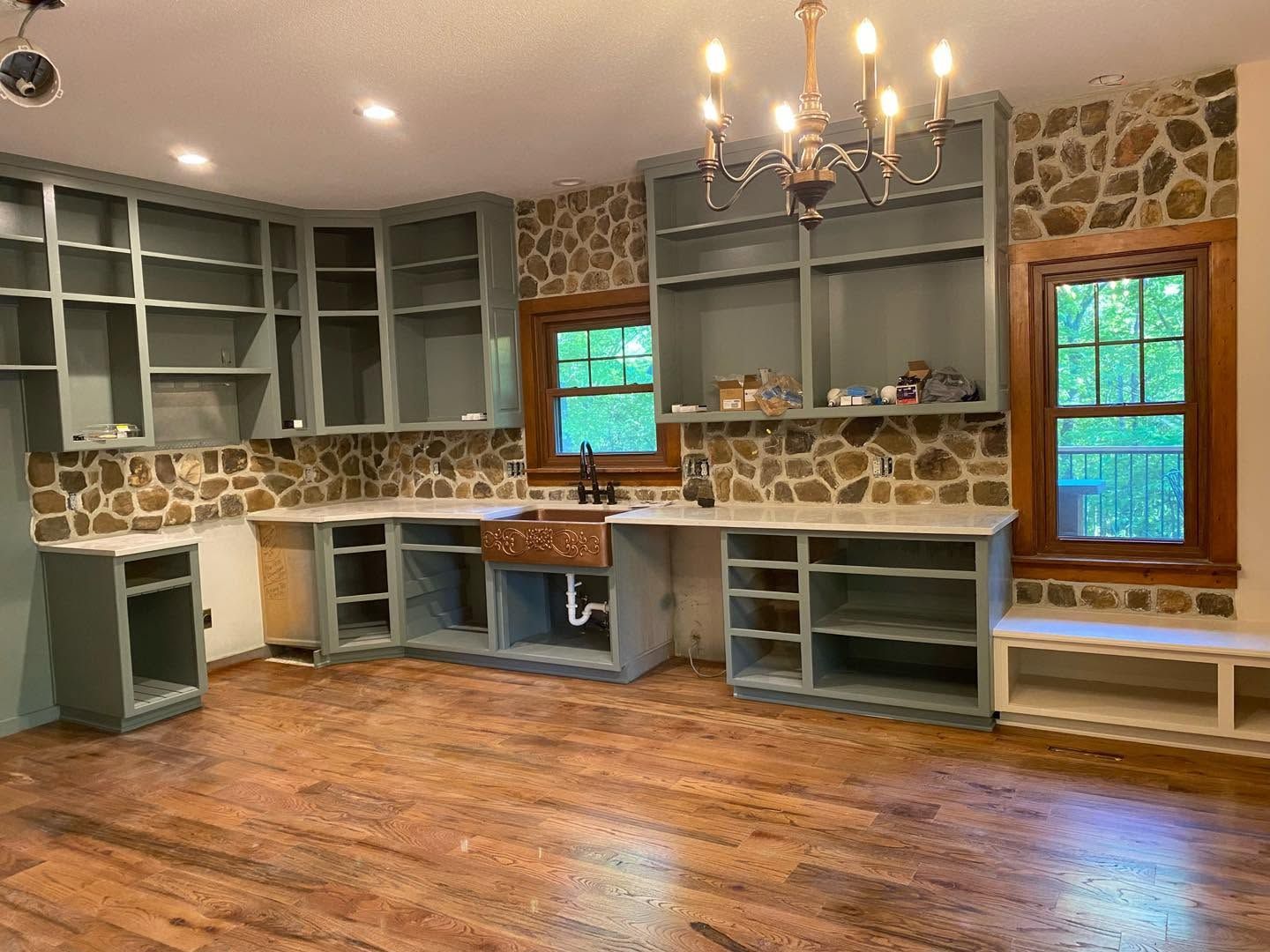 Rustic kitchen with stone backsplash, gray cabinets, wood floor, windows, and chandelier.