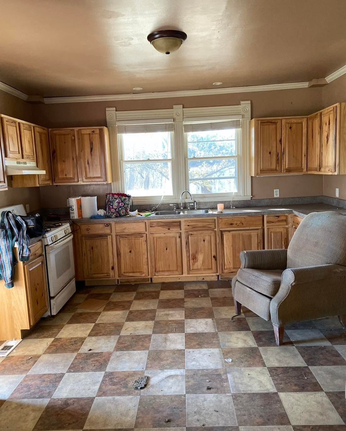 Small rustic kitchen with wooden cabinets, checkered floor, and a gray armchair by the window.