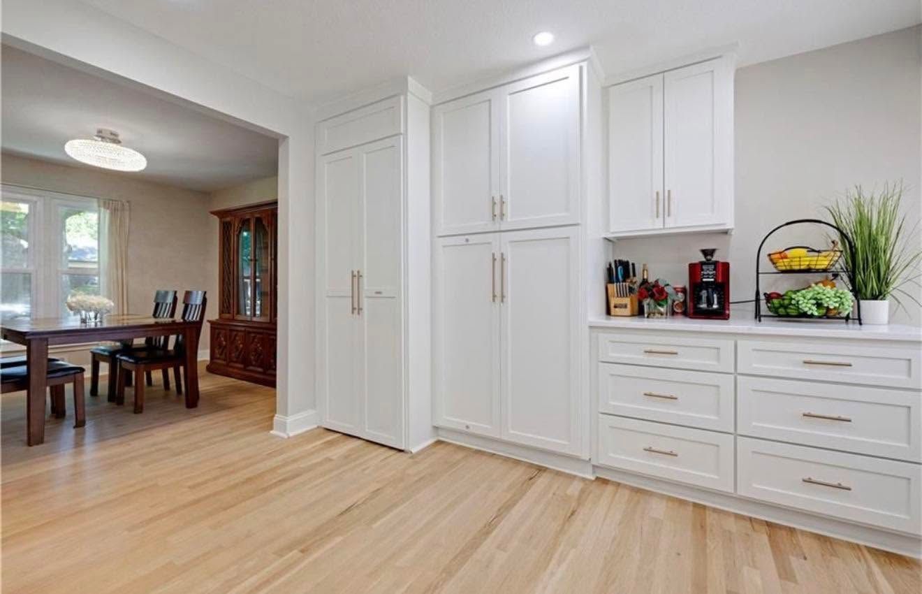 Bright white kitchen with light wood floors, built-in cabinets, and a dining area visible in the background
