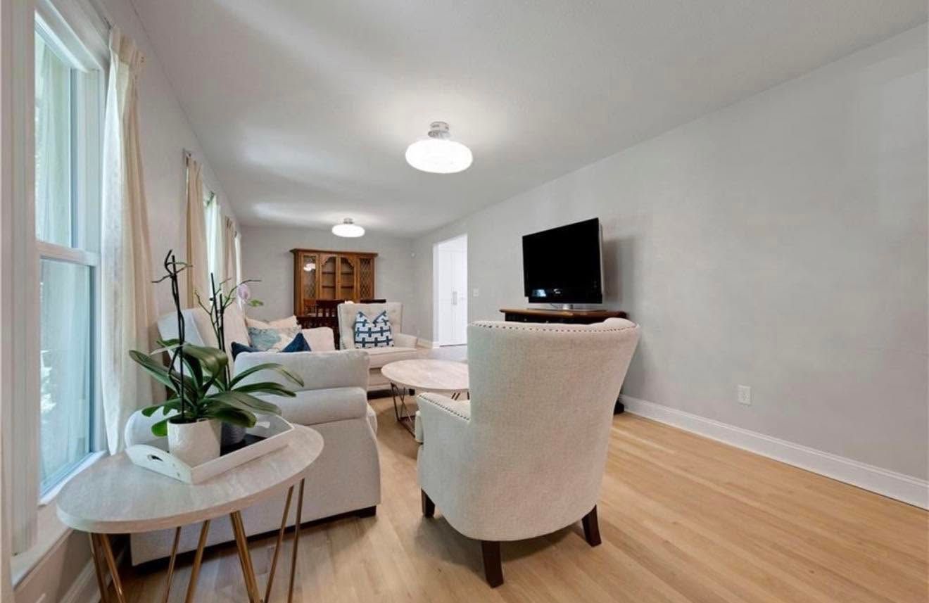 Bright living room with white chairs, wood floors, a TV, and large windows letting in daylight