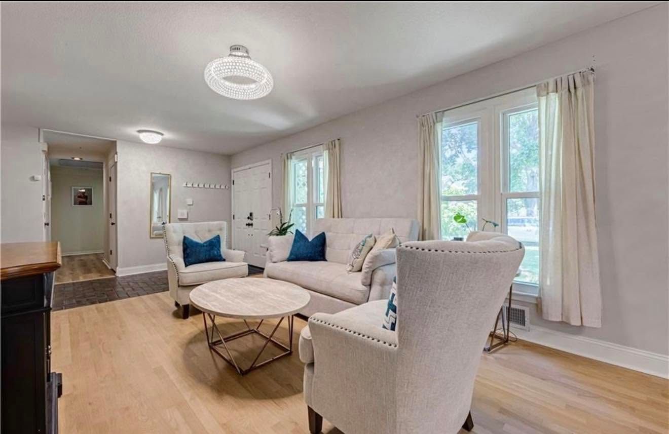 Bright living room with light wood floors, white sofa and armchair, round coffee table, and large windows with curtains