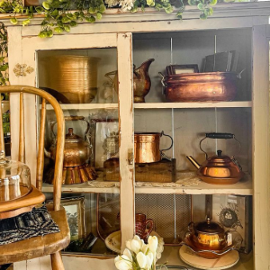 Rustic shelf with copper pots, baskets, greenery, and a wooden chair in a sunlit room
