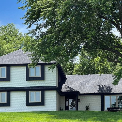 Two-story white house with dark trim under large trees on a green lawn