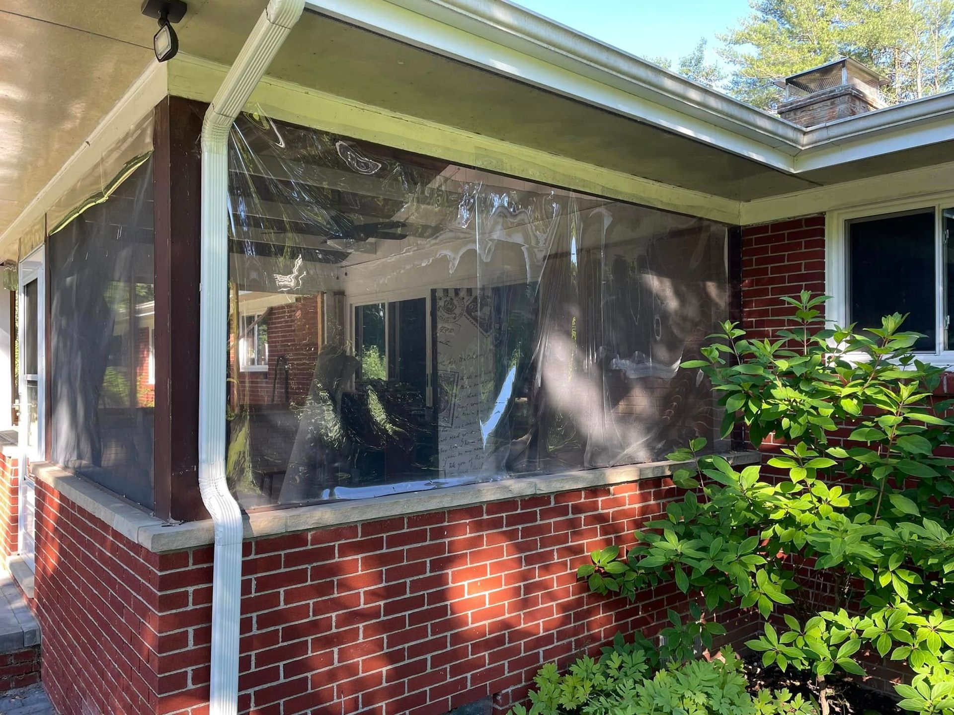 Clear vinyl curtains enclose a brick porch, with a visible gutter and greenery.