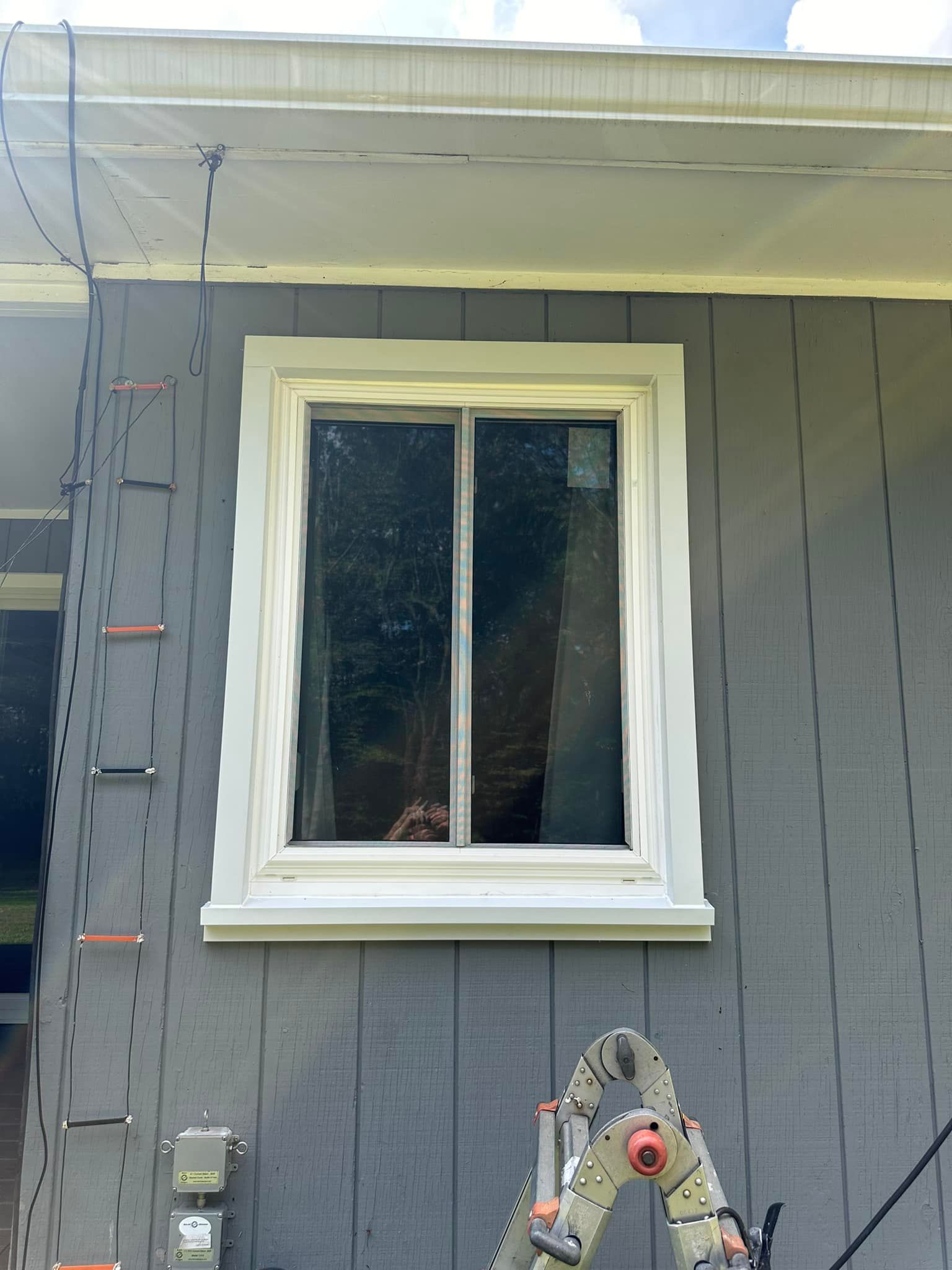Window with white trim on a gray house, ladder in front, under a white eave.