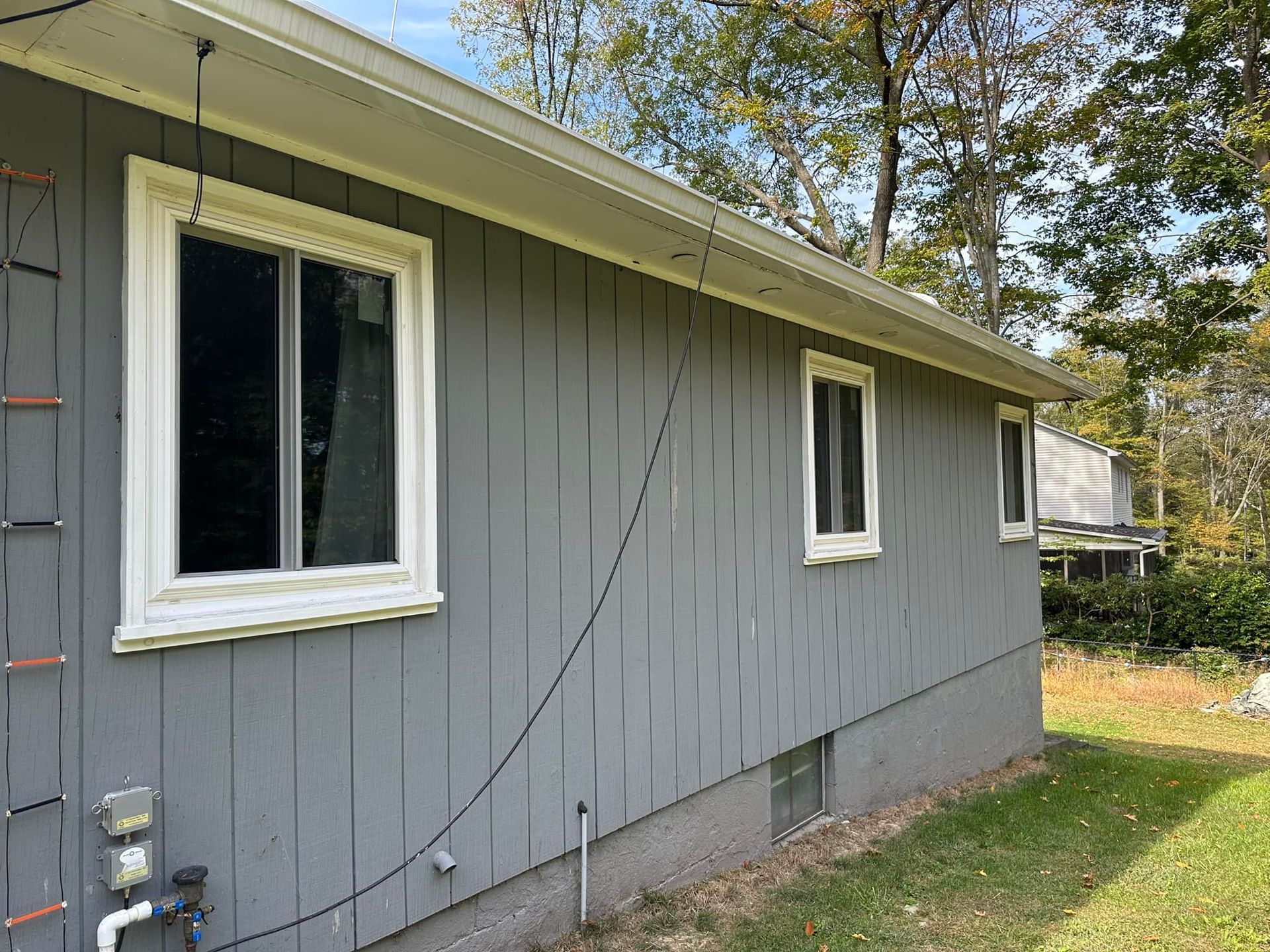 Gray house exterior with white-trimmed windows and a gutter, on a grassy lawn with trees in the background.