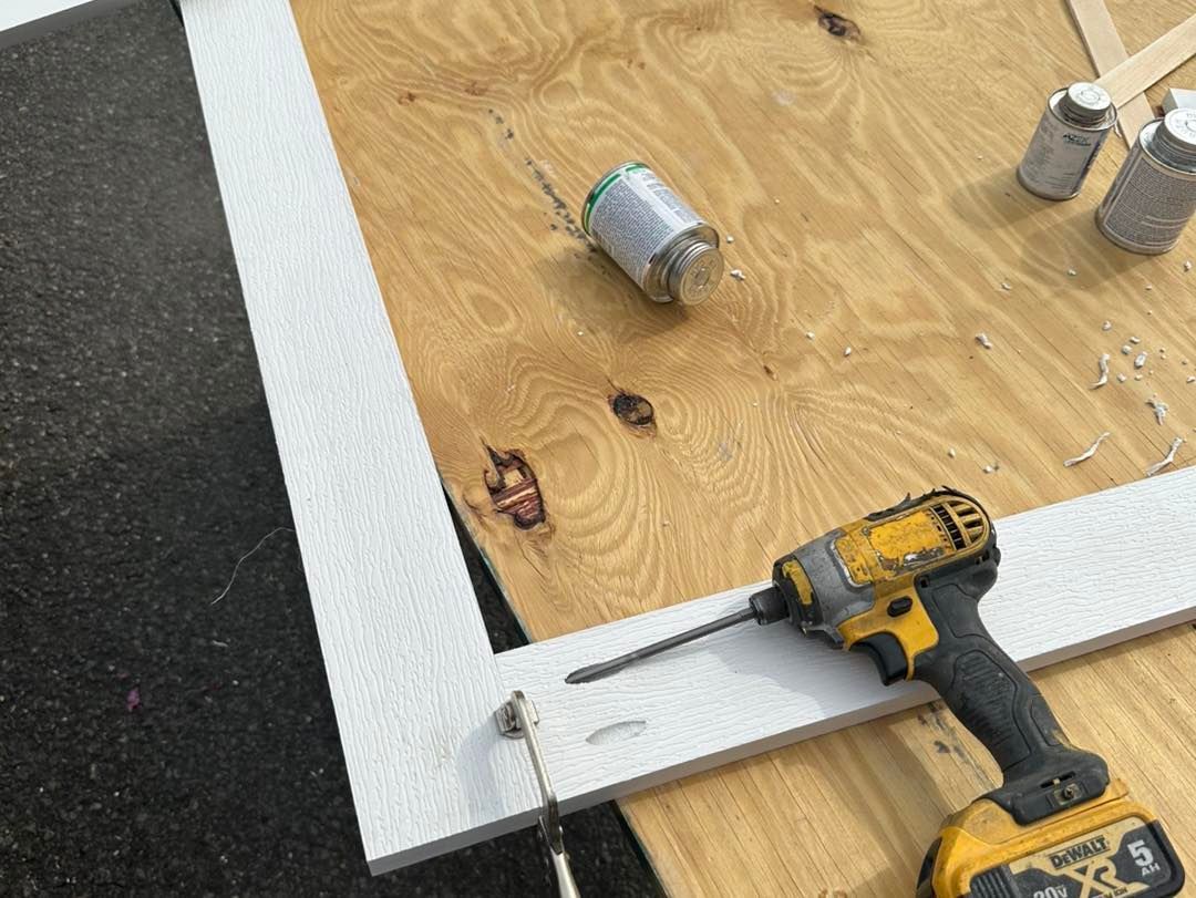 Yellow drill on white wood frame laid on plywood. Silver cans and glue are nearby.