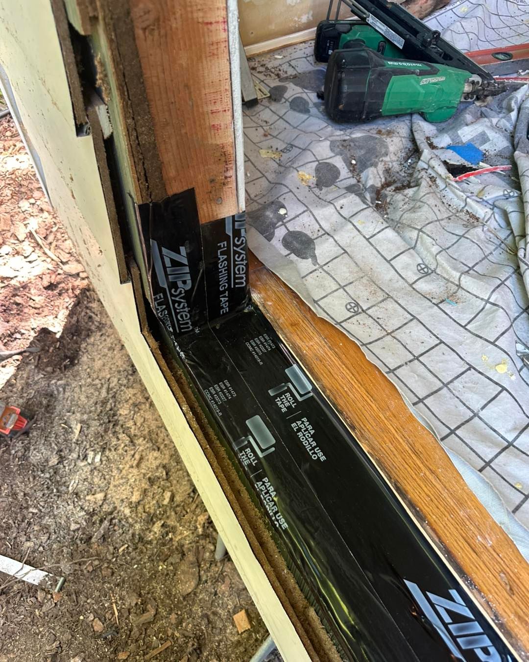 Black flashing tape applied to a window frame corner during construction, green nail gun in the background.