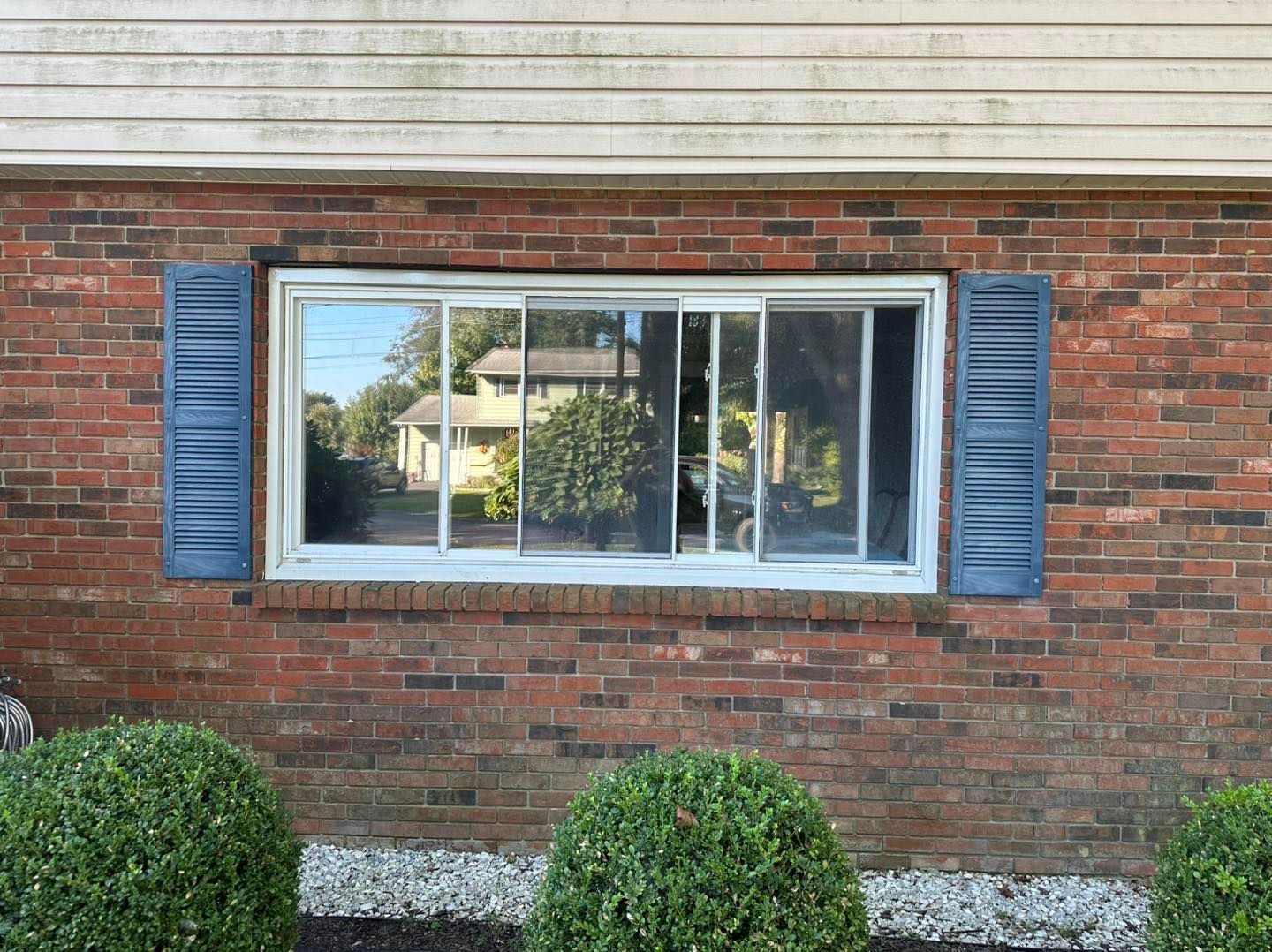 Brick building with a white-framed window. Blue shutters flank the window. Green bushes below.