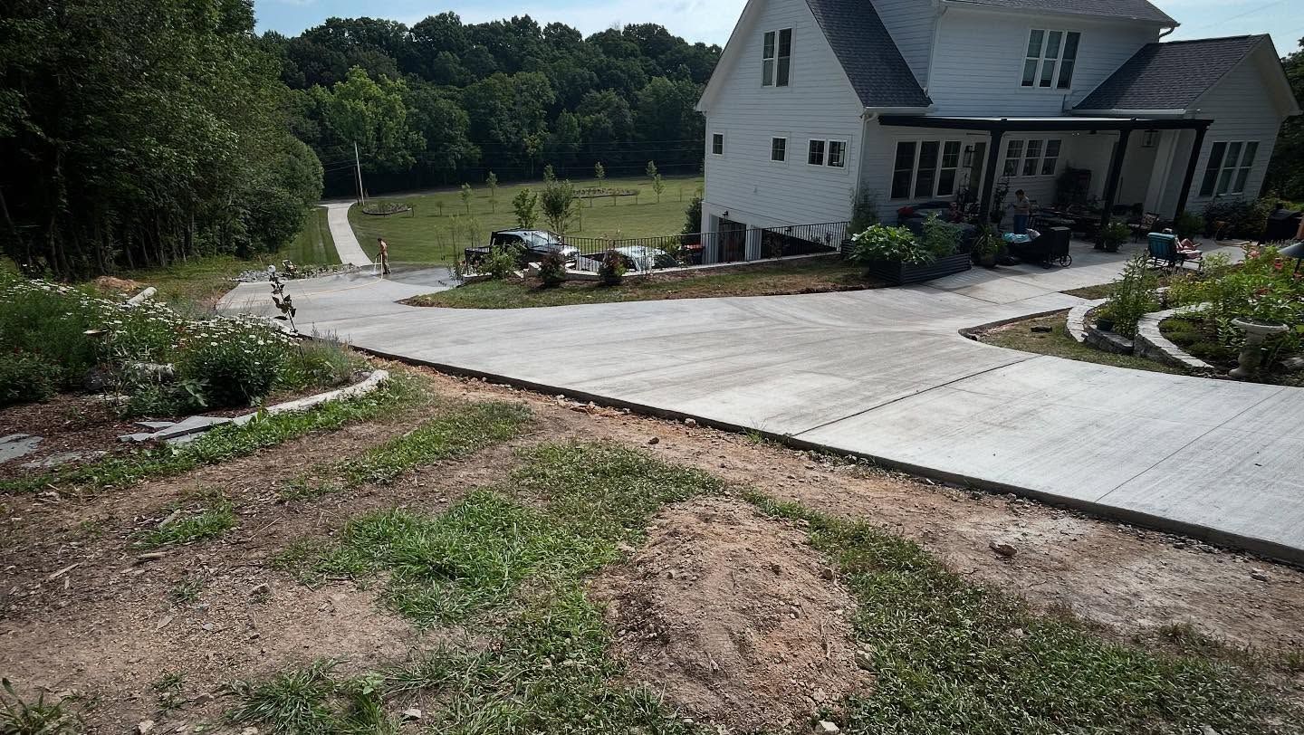 Wide concrete driveway leading to a white house beside a wooded yard and garden beds