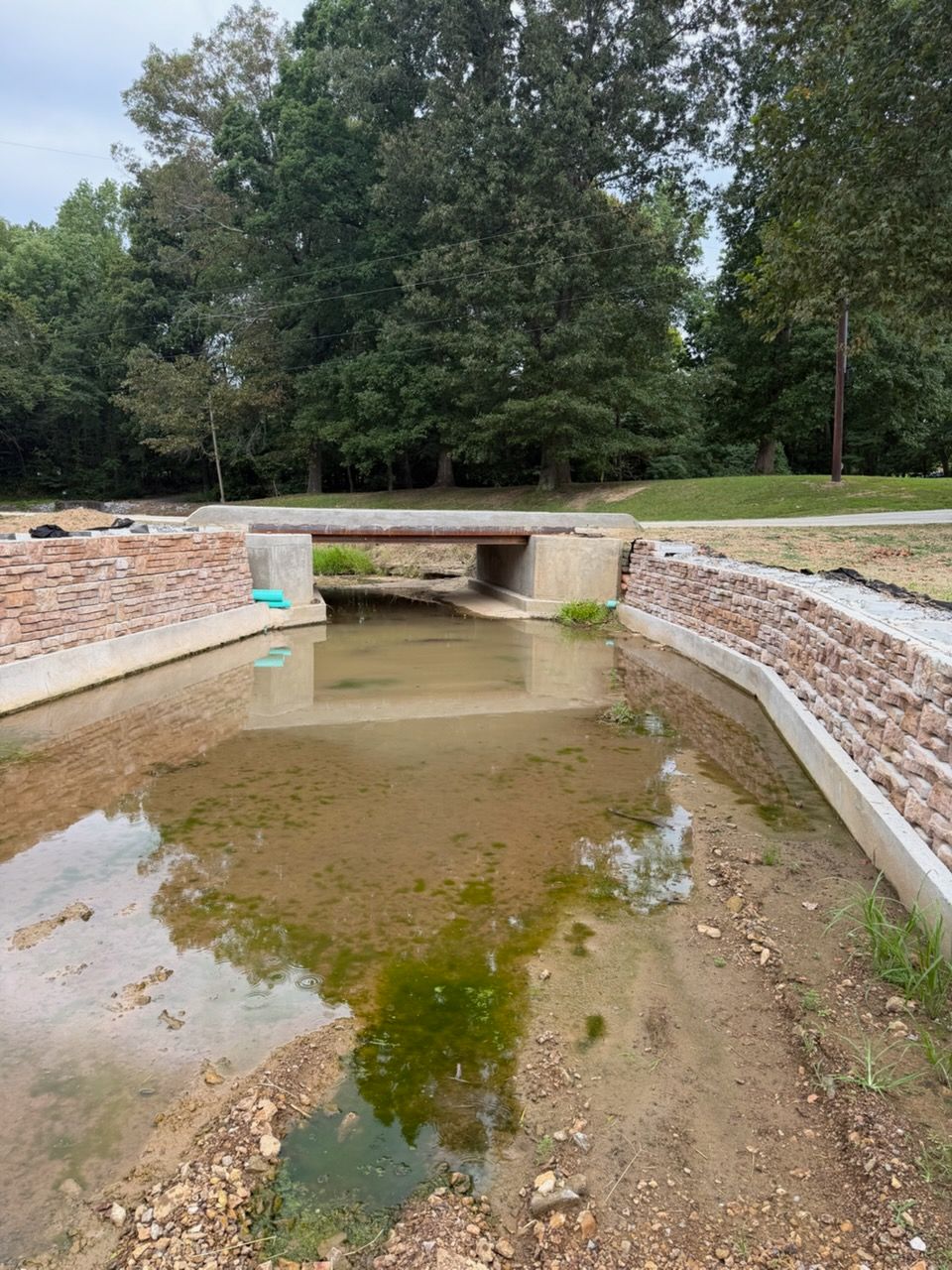 Dry concrete channel with shallow water under a small culvert, lined by stone walls and trees beyond