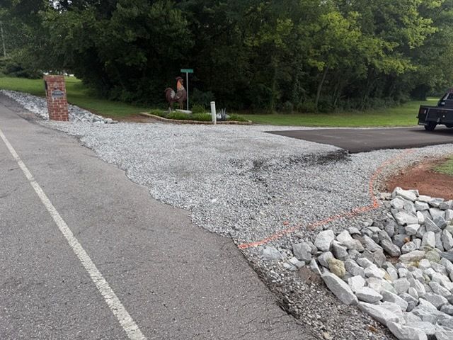 Gravel driveway entrance beside paved road, with rocks, brick pillars, and trees in the background
