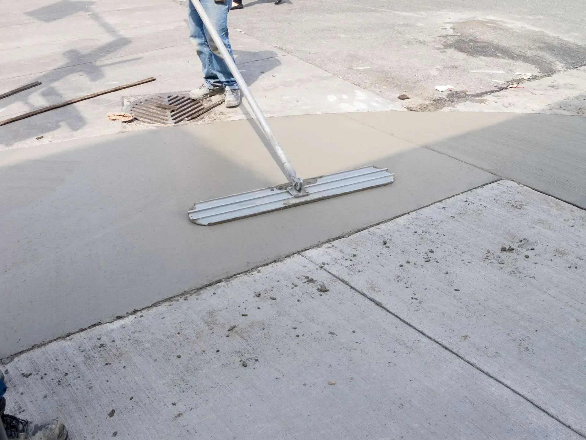 Person pushing a broom over a wet concrete sidewalk near a street drain