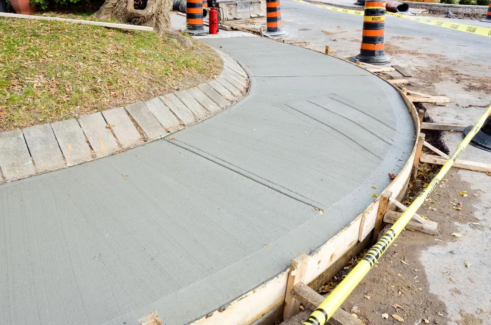 Curved concrete sidewalk under construction beside a road, with caution tape and orange traffic barrels.