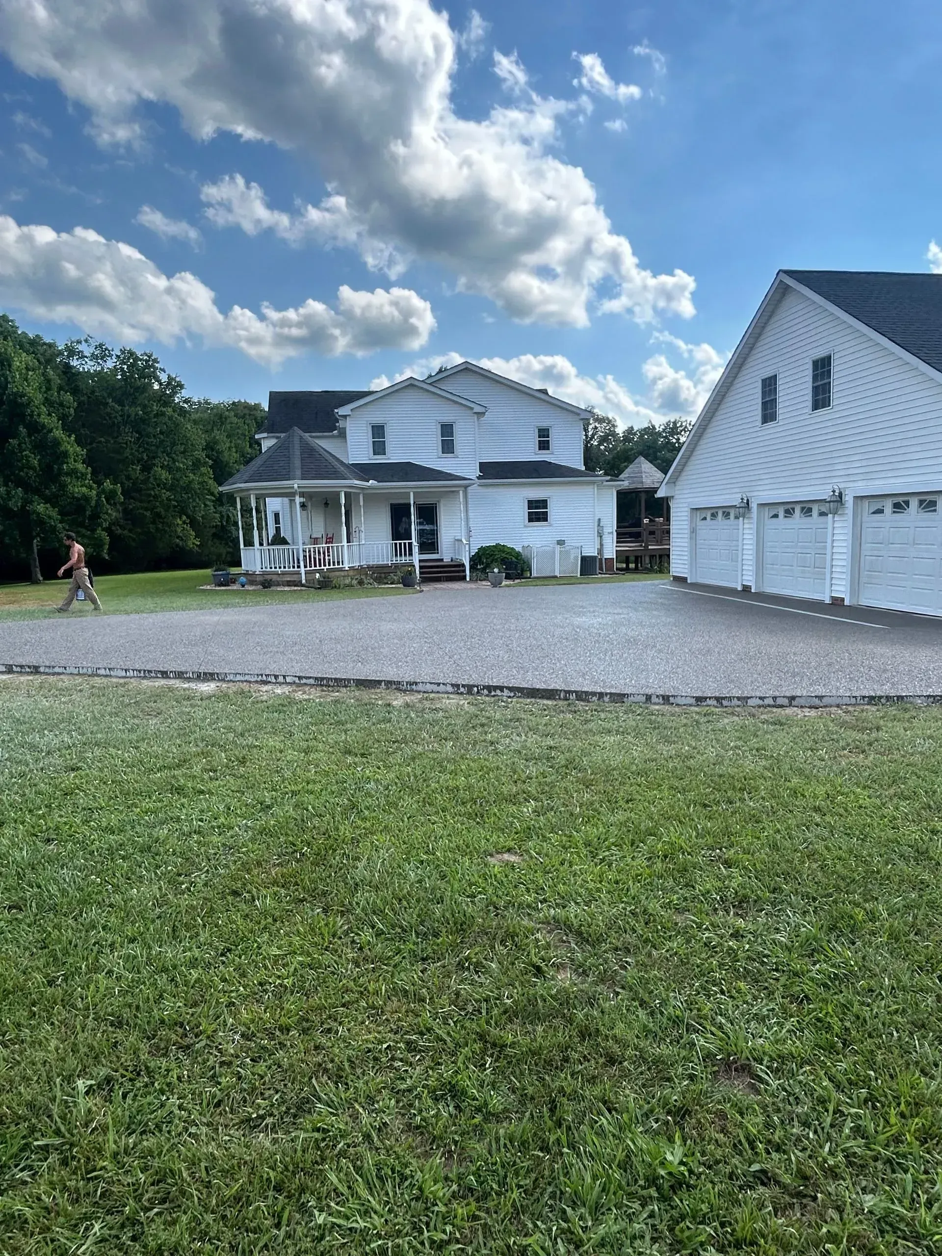 White farmhouse and barn beside a gravel driveway under a cloudy blue sky; a person walks at left.