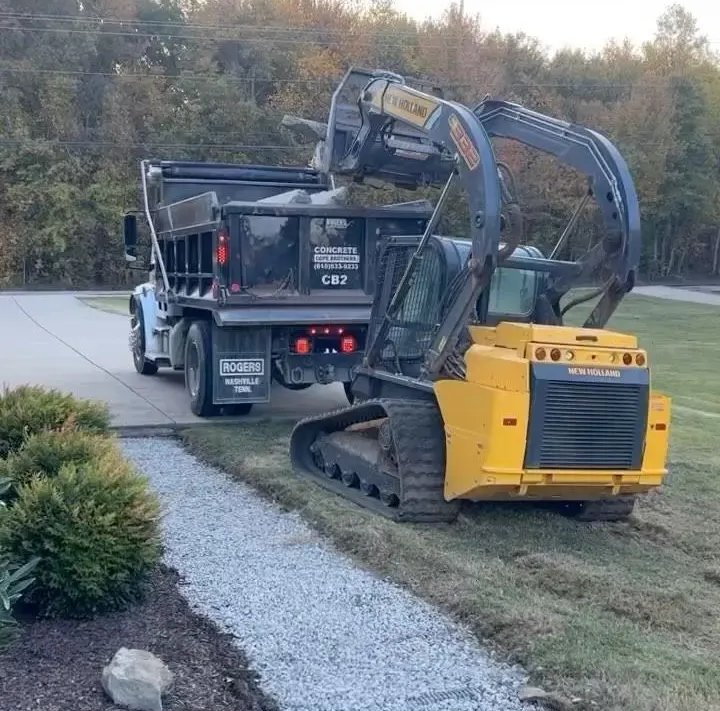 Yellow compact excavator on a trailer beside a gravel driveway, with trees in the background.