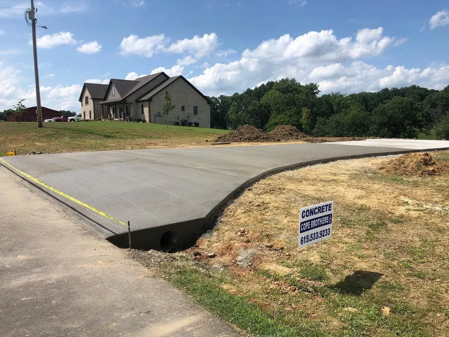 Newly paved road with a drainage gap, near a house and a small sign on a grassy roadside.