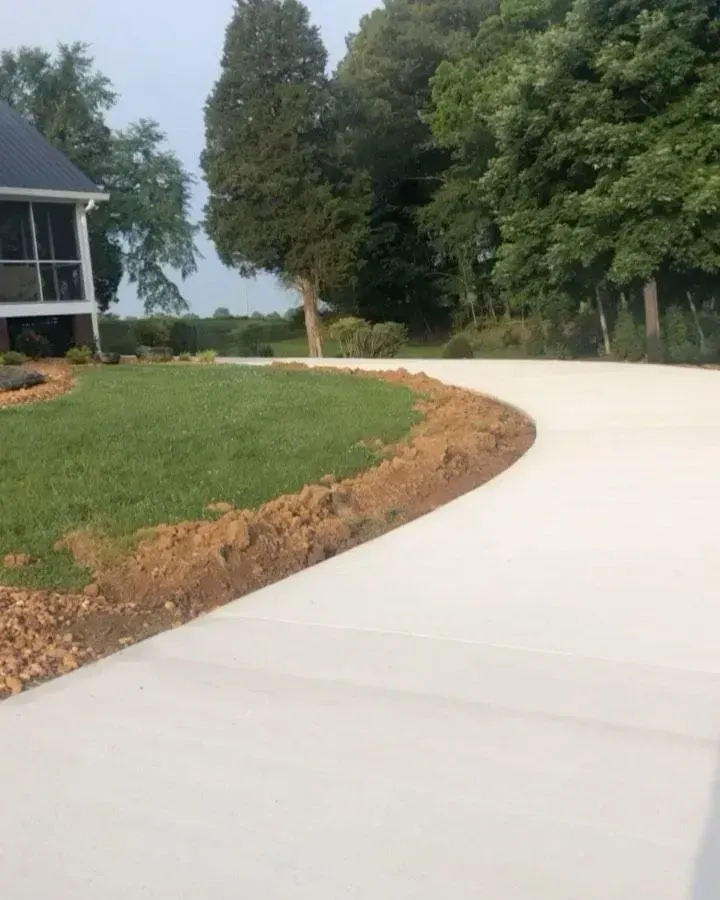 Curving concrete driveway beside a grassy lawn and trees, leading past a house in a wooded setting