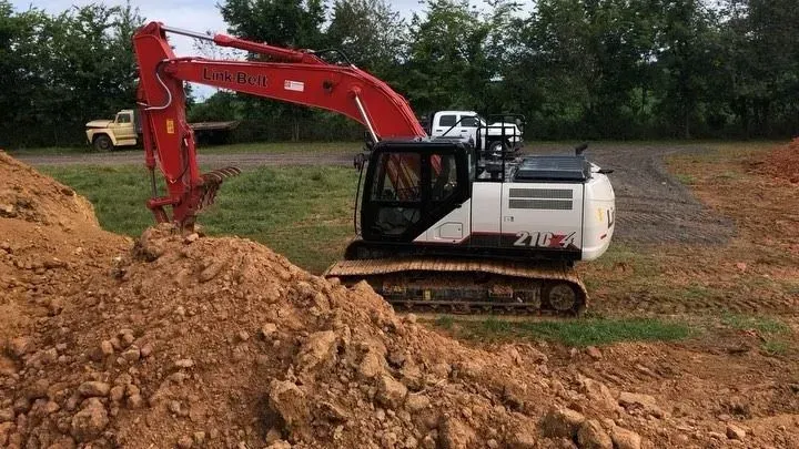Red excavator digging a dirt pile at a construction site, with trees in the background.