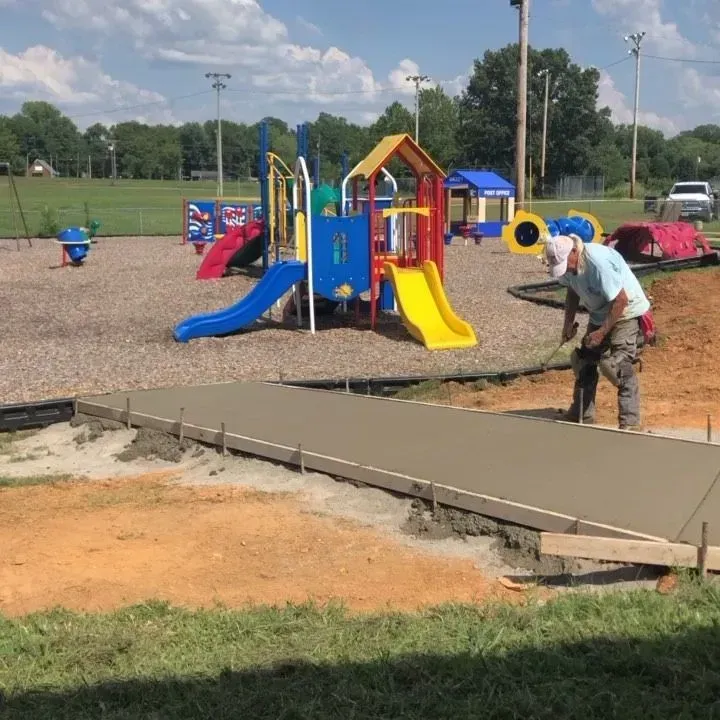 Worker smoothing fresh concrete beside a colorful playground in a park