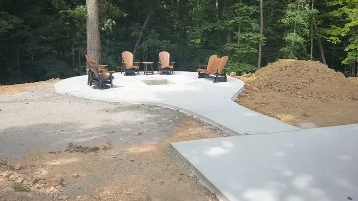Concrete patio foundation with four chairs around a fire pit beside a dirt pile in a wooded yard