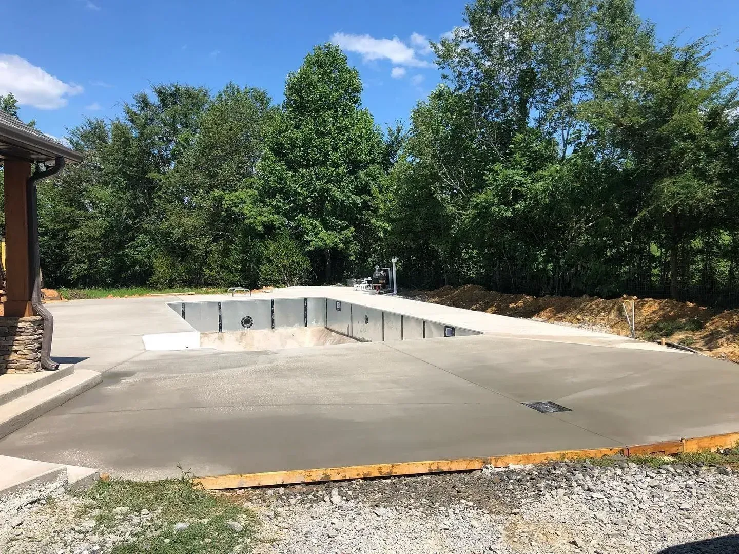 Empty concrete swimming pool shell beside a driveway, surrounded by trees and a house corner.