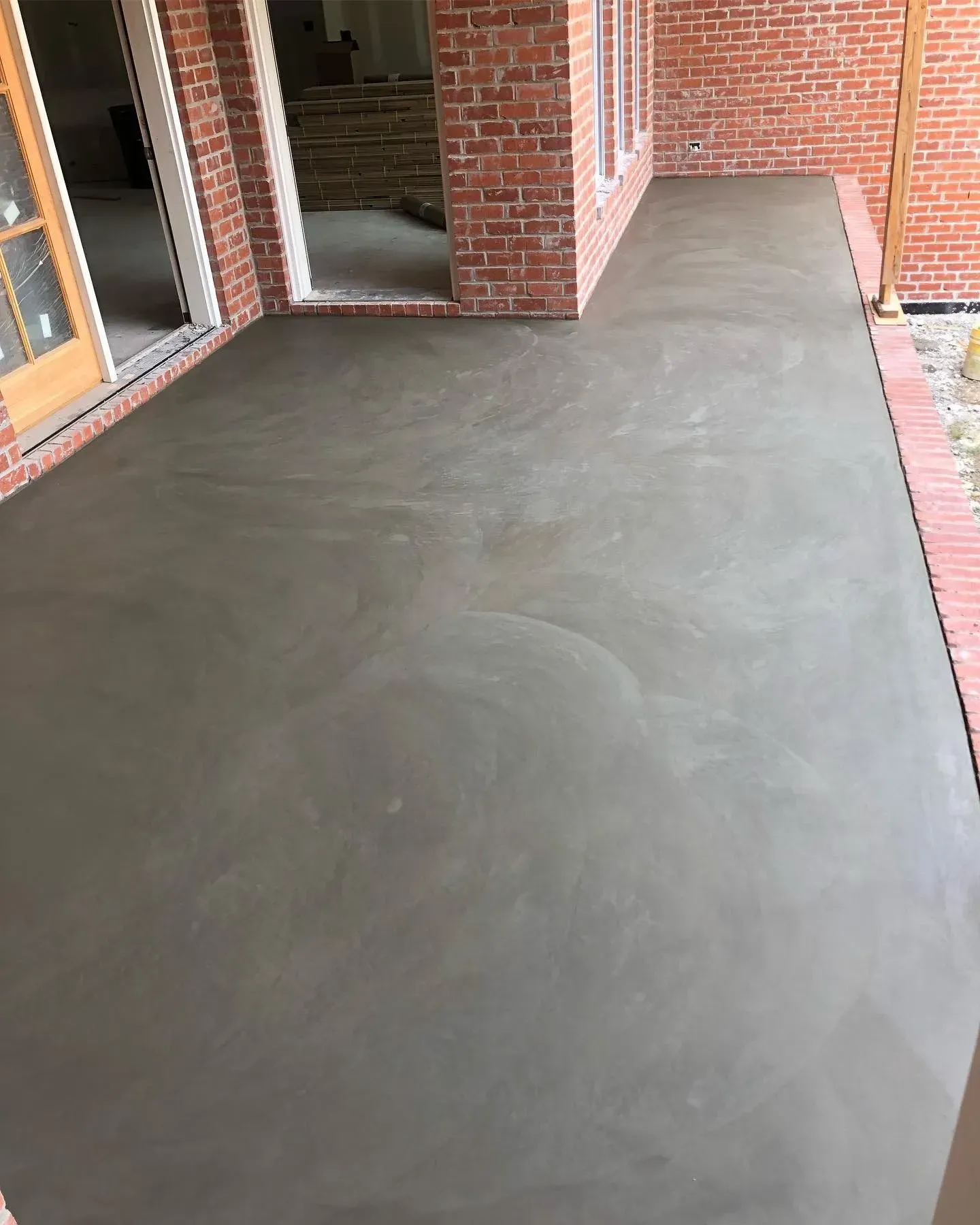 Covered concrete patio beside a brick house, with a smooth gray floor and open doorway.