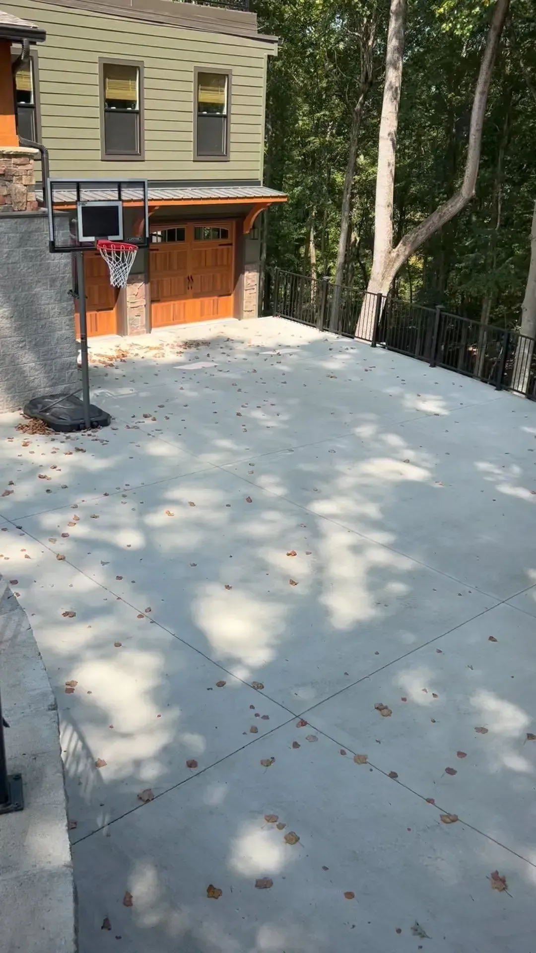 Concrete patio beside a beige house, shaded by trees, with a wooden garage door and wooded slope nearby