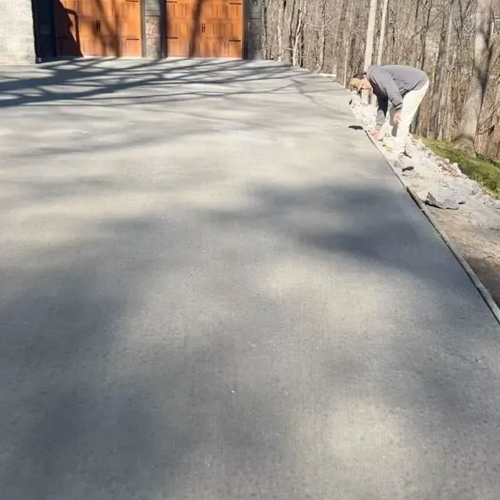Person leveling a concrete driveway beside a wooden fence and gate on a sunny day