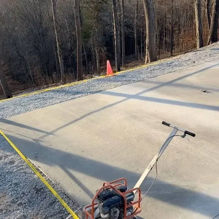 Concrete driveway being smoothed with a power trowel, marked by yellow tape and an orange cone nearby