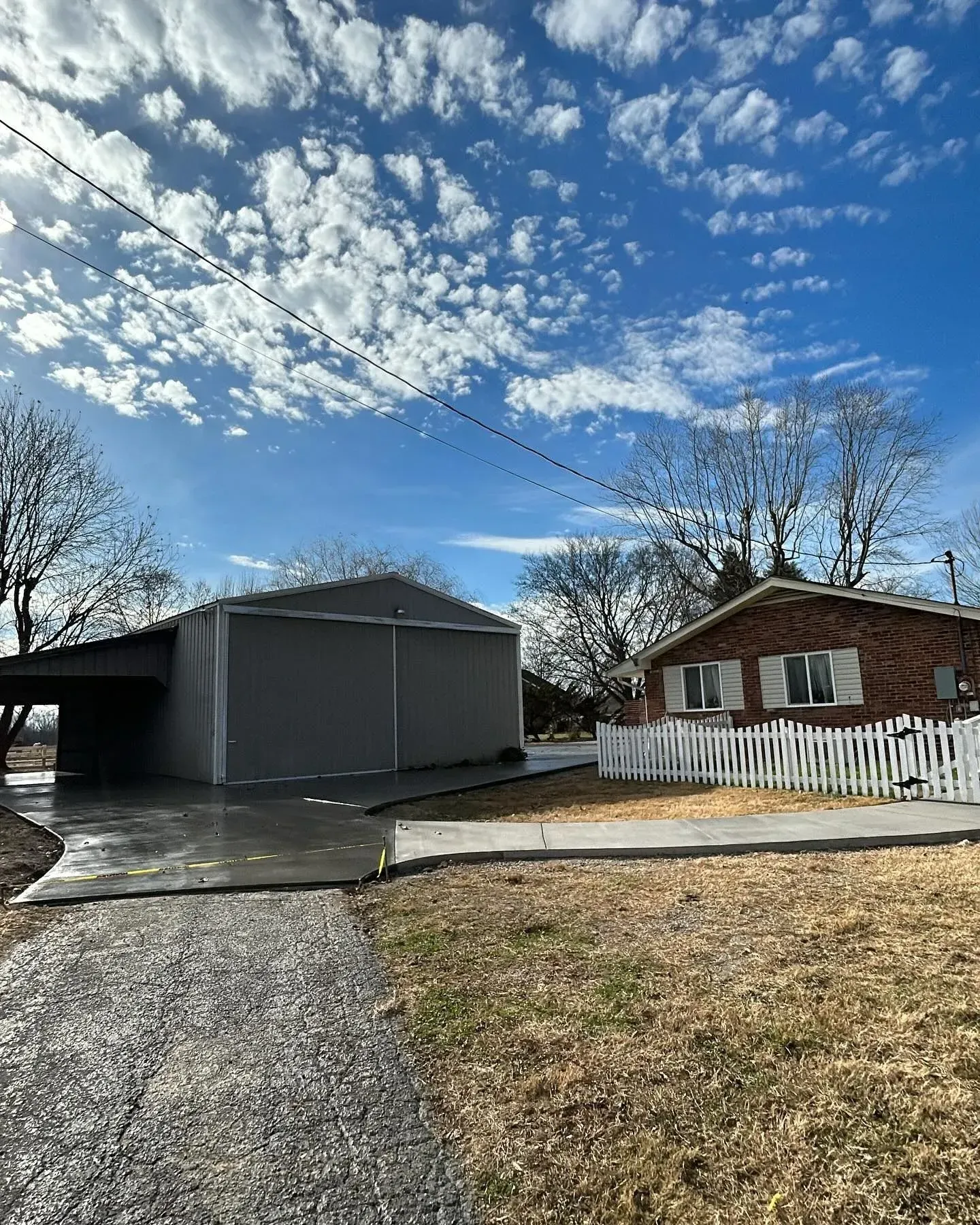 Driveway beside a gray garage and brick house under a bright, cloud-streaked sky
