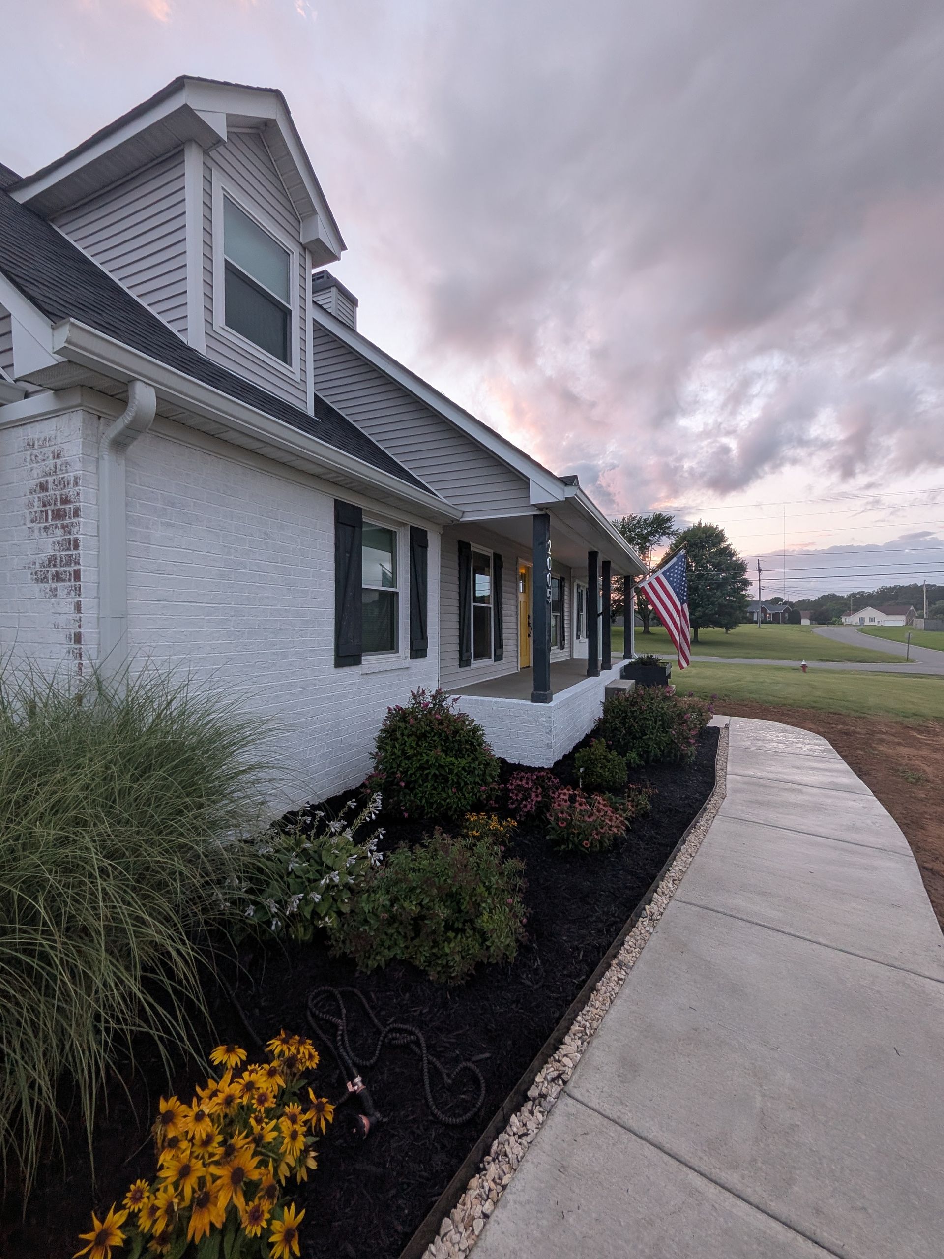 Driveway beside a gray house with a brick patio, lawn, and trees in the background.