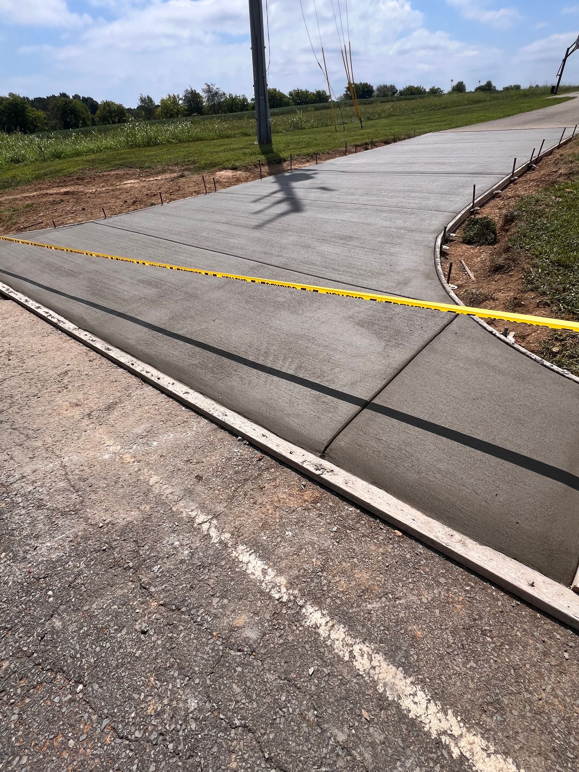 Caution tape blocking a cracked concrete path beside a road and grassy field.